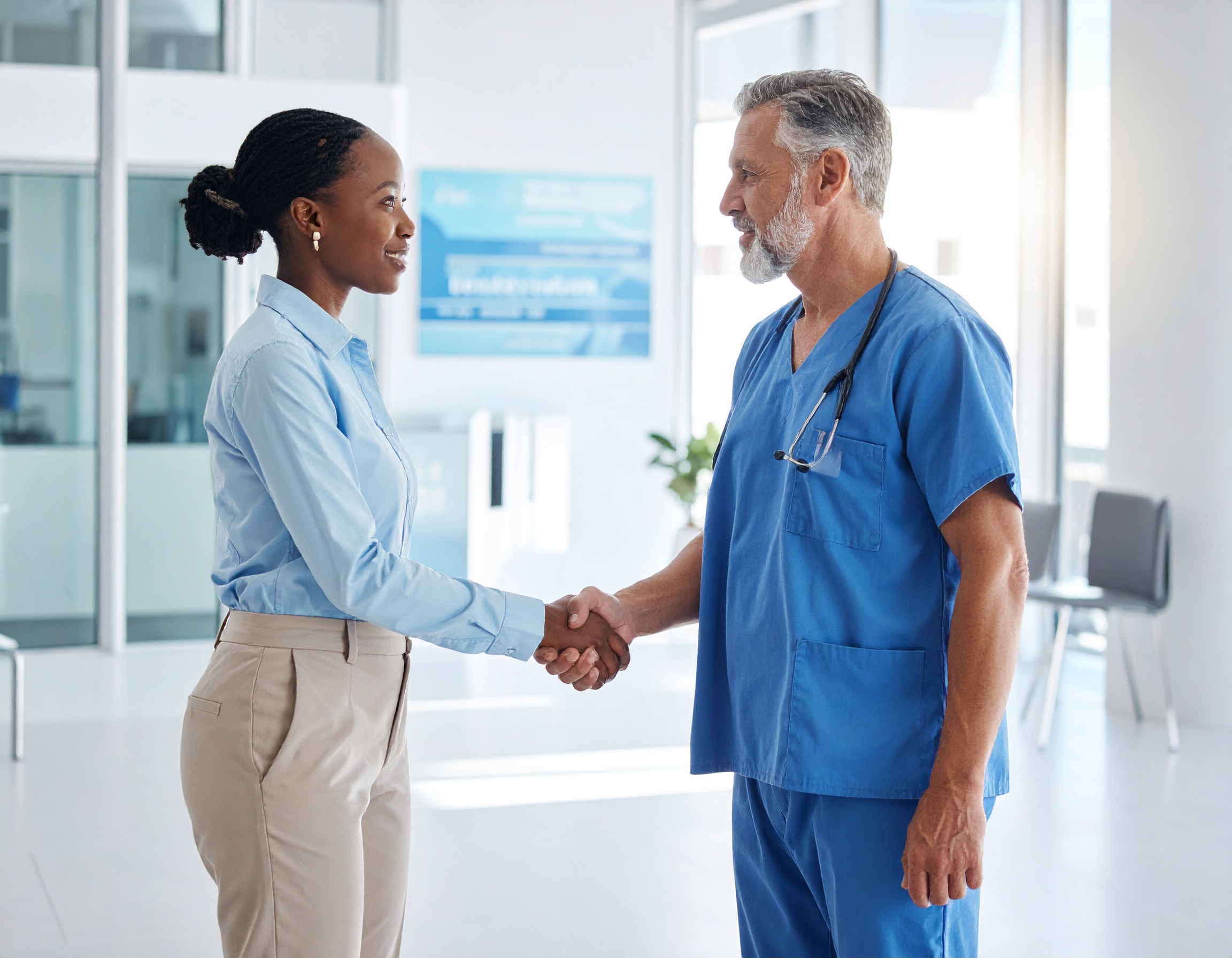 Younger professional shaking hands with a senior consultant in blue scrubs in a hospital during a pre-interview visit.