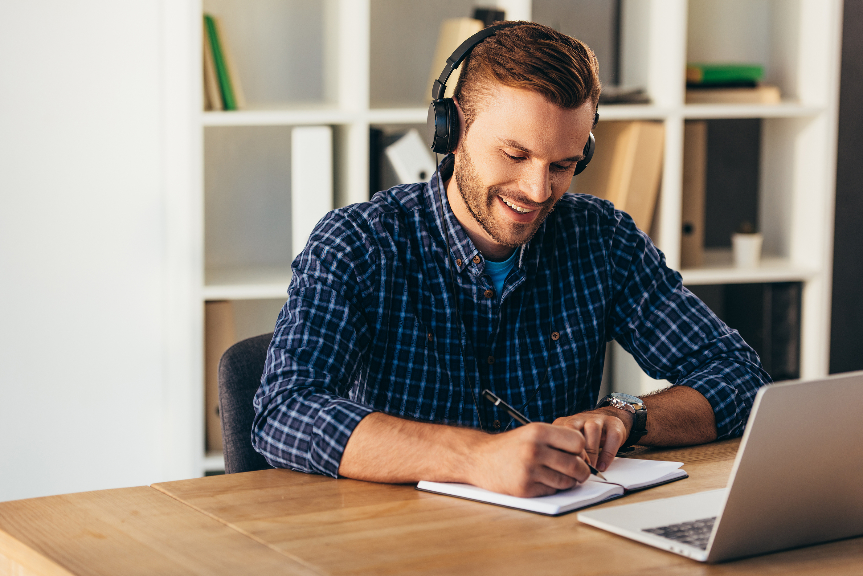 Stock image of a smiling professional wearing headphones, taking notes at a laptop, representing structured consultant interview preparation and use of question bank.