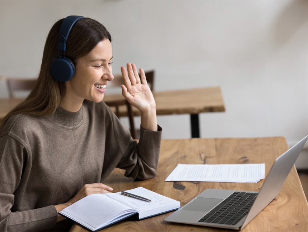 A friendly woman with headphones waves at her laptop, participating in an online NHS consultant interview skills coaching session.